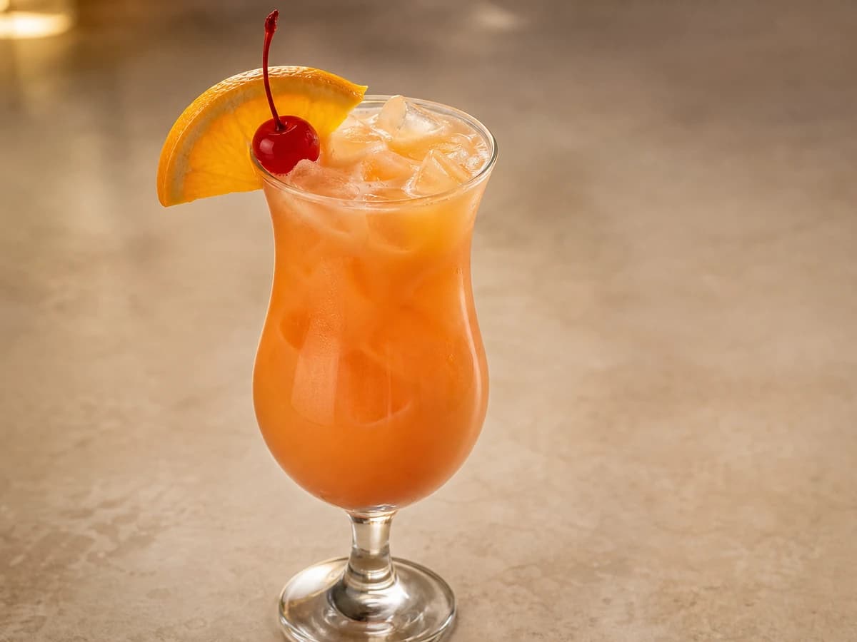 A Bahama Mama in a hurricane glass with an orange slice and cherry, showing a sunset gradient of colour