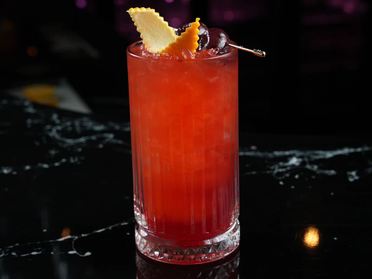 A tall, reddish-pink Singapore Sling in a hurricane glass garnished with pineapple and a cherry